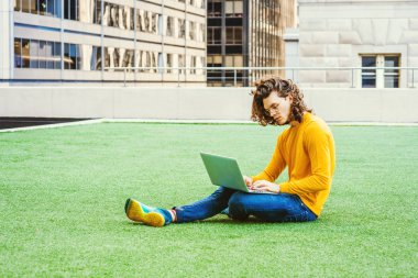New York 'ta okuyan, kahverengi kıvırcık saçlı, gözlüklü, sarı uzun kollu tişörtlü, kot pantolonlu, spor ayakkabılı yeşil çimlerde oturan, dizüstü bilgisayarında çalışan İspanyol asıllı genç bir üniversite öğrencisi..