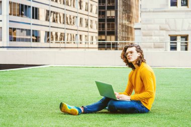New York 'ta okuyan, kahverengi kıvırcık saçlı, gözlüklü, sarı uzun kollu tişörtlü, kot pantolonlu, spor ayakkabılı yeşil çimlerde oturan, dizüstü bilgisayarında çalışan İspanyol asıllı genç bir üniversite öğrencisi..