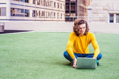 Genç İspanyol Amerikan Koleji Öğrenci sarı uzun kollu T gömlek, yeşil çim üzerinde otururken bacak bacak gözlük, eğitim uzun kıvırcık kumral, New York'ta dizüstü bilgisayar üzerinde çalışan