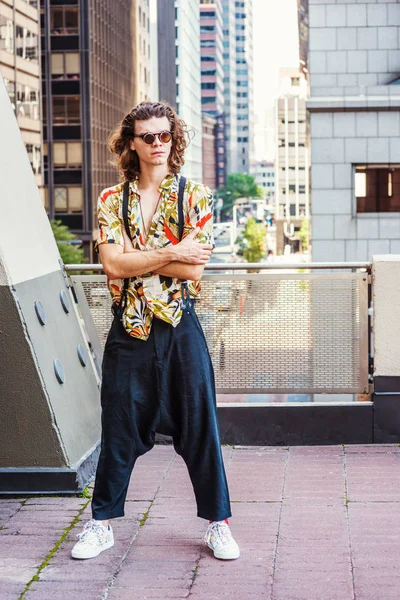 Young Man with brown curly hair, wearing sunglasses, patterned short sleeve shirt, baggy loose pants with suspenders, patterned sneakers, hanging old key as necklace, standing on balcony in New York