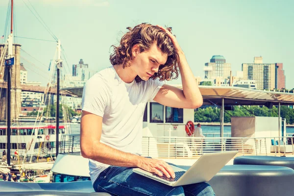 Genç İspanyol Amerikan seyahat, New York, uzun kıvırcık saçları ile çalışan adam beyaz T gömlek, kot, Doğu ırmak, dizüstü bilgisayarda çalışarak oturan el saç dokunuyor, düşünme