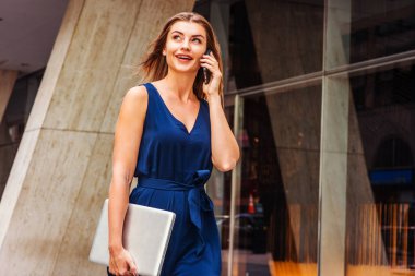 Doğu Avrupalı genç bir kadın cep telefonuyla konuşuyor, seyahat ediyor, New York 'ta çalışıyor, mavi kolsuz tulum giyiyor, dizüstü bilgisayar taşıyor, ofis binasının dışında sokakta yürüyor.