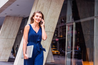Doğu Avrupalı genç bir kadın cep telefonuyla konuşuyor, seyahat ediyor, New York 'ta çalışıyor, mavi kolsuz tulum giyiyor, dizüstü bilgisayar taşıyor, ofis binasının dışında sokakta yürüyor.