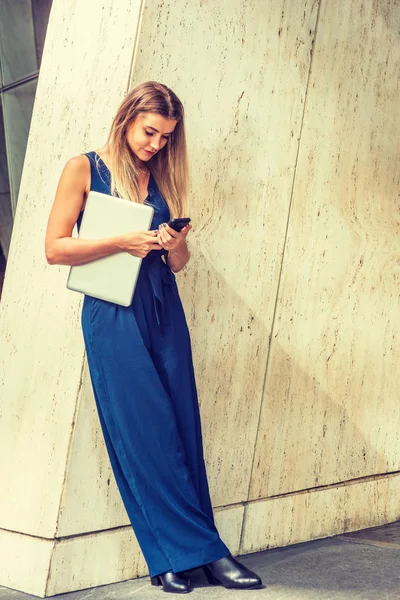 東ヨーロッパ アメリカで若い女性長い茶色の髪 青いノースリーブ ジャンプ スーツ 黒革の靴を履いて ニューヨーク 携帯電話のテキスト メッセージの列に対して立っているラップトップ コンピューターを保持 — ストック写真