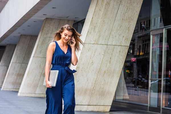 Young Eastern European American Woman talking on cell phone, traveling, working in New York City, wearing blue sleeveless jumpsuit, carrying laptop computer, walking on street outside office building