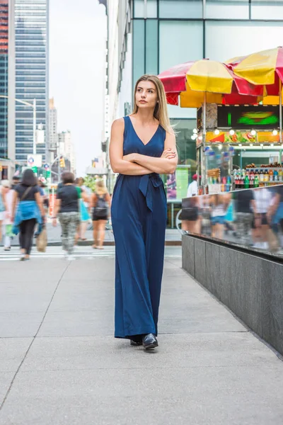 Young Eastern European Woman traveling in New York, wearing blue sleeveless jumpsuit, black leather shoes, crossing arms, looking around,  walking on busy street in middletown of Manhattan