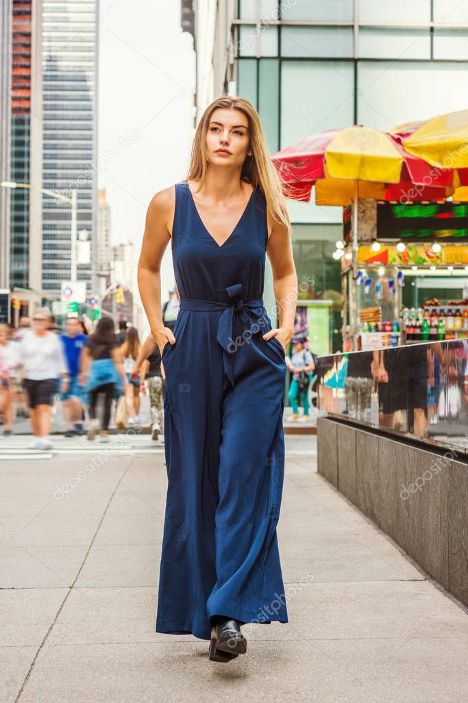 Young Eastern European Woman with long brawn hair traveling in New York City, wearing blue sleeveless, v neck, jumpsuit, black leather shoes, walking on busy street in middletown of Manhattan