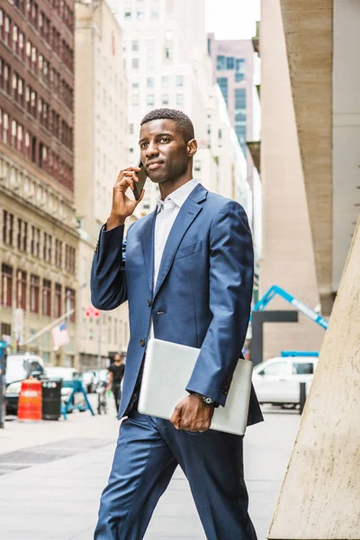 Genç Afrikalı Amerikalı iş adamı cep telefonuyla konuşuyor, seyahat ediyor, New York 'ta çalışıyor, mavi takım elbise giyiyor, beyaz atlet giyiyor, dizüstü bilgisayar taşıyor, ofis binasından caddeye yürüyor..