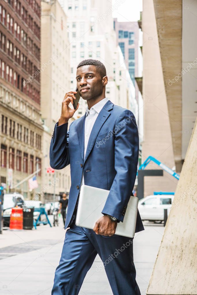 Young African American businessman talking on cell phone, traveling, working in New York, wearing blue suit, white undershirt, carrying laptop computer, walking out from office building to street.