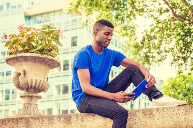 Modern okuma. Genç Afro-Amerikan üniversite öğrencisi New York'ta giyiyor mavi T gömlek, siyah pantolon, spor ayakkabı, kampüste, duvar üzerine oturan, seyir, okuma mavi tablet bilgisayar eğitim