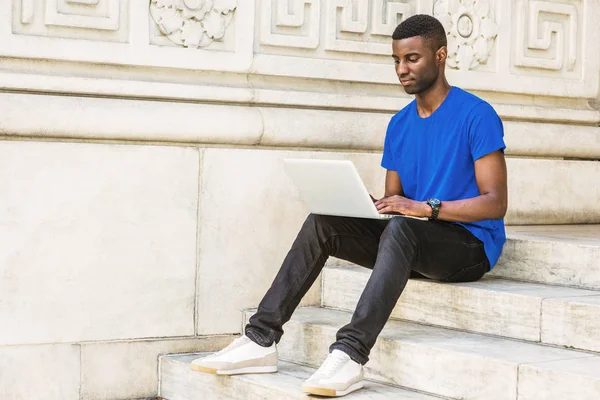 New York 'ta okuyan, mavi tişörtlü, siyah pantolonlu, spor ayakkabılı, kol saati takan, ofis binasının dışındaki merdivenlerde oturan, kampüste dizüstü bilgisayarla çalışan genç bir Afrikalı Amerikalı.