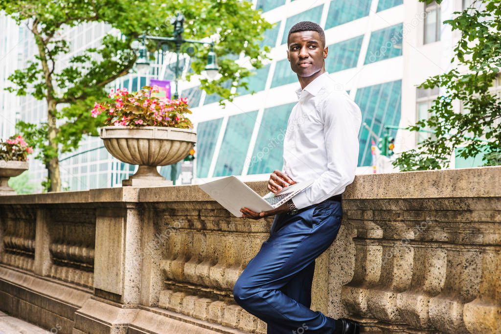 Young African American College Student studying, working in New York, wearing white long sleeve shirt, blue pants, standing against half vintage wall on campus, holding laptop computer, looking away