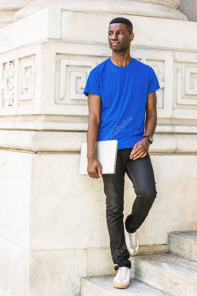 Young African American Graduate Student studying in New York, wearing blue T shirt, black pants, sneakers, wristwatch, holding laptop computer, standing on stairs outside office building on campus