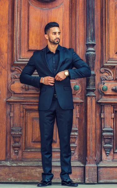 Portrait of Young East Indian American Businessman with beard in New York, wearing black suit, leather shoes, wristwatch, standing by brown old style office doorway, hands buttoning jacket