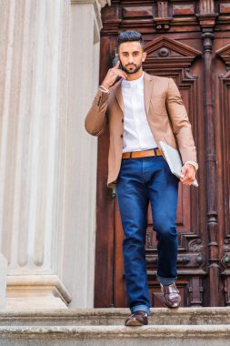 Genç Doğu Hint Amerikalı kahverengi ceket, beyaz giyen işadamı sakallı, yuvarlak yakalı gömlek, pantolon, dizüstü bilgisayar, tutarak merdivenden office tarafından kapı gelirken yürümeye cep telefonuyla konuşurken mavi