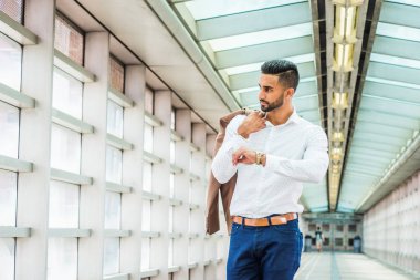 Genç Doğu Hint Amerikalı ceket omzunun üzerinden taşıyan beyaz gömlek giymiş işadamı New York'ta sakallı içinde kapalı geçit ayakta, kol kol saati ile yükselterek, uzağa arıyorsunuz, düşünme