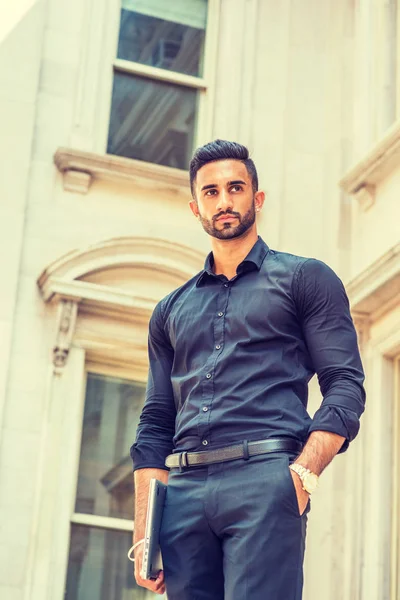 Portrait of Young East Indian American Businessman with beard in New York, wearing black long sleeve shirt, black pants, holding laptop computer, standing outside old style office building, looking u