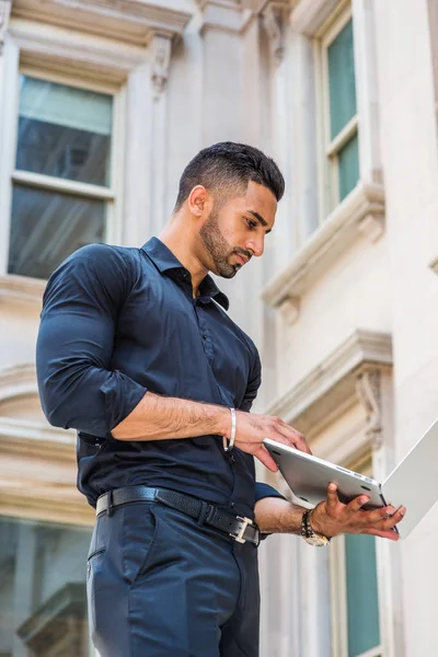 Way to Success. Young East Indian American Businessman with beard working in New York, wearing black shirt, holding laptop computer, standing outside old style office building, looking down, reading