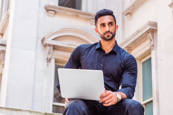 Way to Success. Young East Indian American Businessman with beard working in New York, wearing black shirt, holding laptop computer, sitting outside old style office building, looking up, thinking