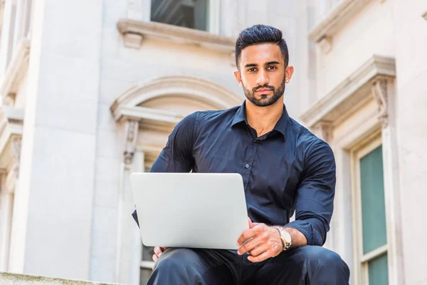 Way to Success. Young East Indian American Businessman with beard working in New York, wearing black shirt, holding laptop computer, sitting outside old style office building, looking, thinking