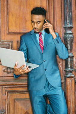New York'ta, gök mavi takım giyen, çalışma sakallı genç Afrikalı-Amerikalı işadamı mor kırmızı desenli kravat, dizüstü bilgisayar, cep telefonuyla konuşurken üzerinde kahverengi office kapının yanında duran