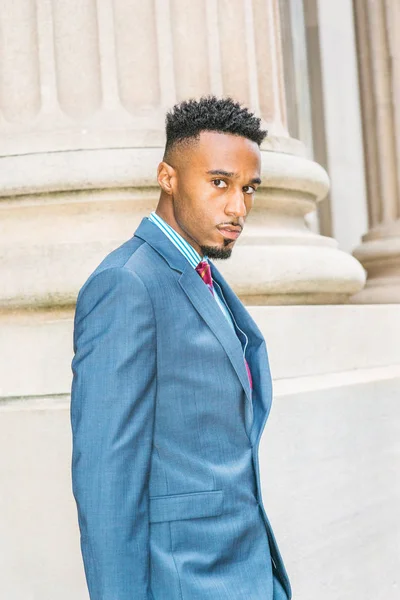 After work. Young African American Businessman with beard, short afro hair, working in New York, wearing dark sky blue suit, violet red patterned tie, walking out from office building, looking away