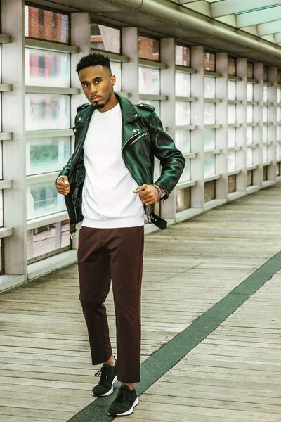 Young Man Casual Fashion in New York. Young African American Guy with beard, wearing black leather jacket, white shirt, black pants, sneakers, standing on walk way with glass wall and wooden floor