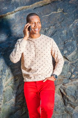 Telefonda konuşan adam. Kol saati, Genç bir siyah adam kayalar tarafından duruyor gülümseyerek, cep telefonundan bir telefon görüşmesi giydiği krem, desenli, yakasız kazak, kırmızı pantolon içinde giyinme.
