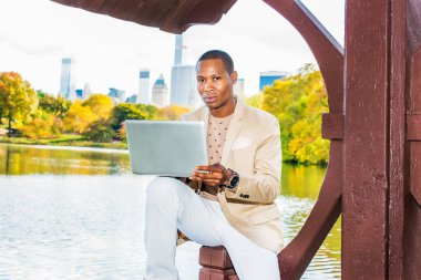 İş adamı çalışma. Giyinme krem blazer, yakasız kazak, beyaz pantolon, bir kol saati, giyen genç bir siyah adam büyük bir şehirde bir gölün yanında oturuyor düşünme, dizüstü bilgisayar üzerinde çalışan.