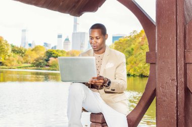 İş adamı çalışma. Krem blazer giyinme, yakasız kazak, beyaz pantolon giyen bir kol saati, siyah adam büyük bir şehirde bir gölün yanında oturan bir genç, aşağı, dizüstü bilgisayarda çalışmak can.