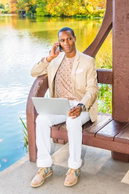 İş adamı dışında. Giyinme krem blazer, yakasız kazak, beyaz pantolon, spor ayakkabı, Genç bir siyah adam bir göl kenarında otururken, telefonda konuşurken, uzağa arıyorsunuz, dizüstü bilgisayar üzerinde çalışan.