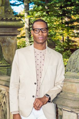 Genç siyah Profesör portresi. Krem blazer giyinme, yakasız kazak giyen gözüne gözlük, kol saati, Genç bir siyah yüksek lisans öğrencisi, kampüste, cidden sana bakıp duruyor.