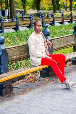 Adam seni bekliyor. Giyinme krem blazer, yakasız kazak, kırmızı pantolon, spor ayakkabı, Genç bir siyah adam parkta bankta oturan, sana bakarken, rahatlatıcı.