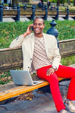 Dışında çalışan adam. Giyinme krem blazer, yakasız kazak, kırmızı pantolon, spor ayakkabı, Genç bir siyah adam bankta oturuyor gülümseyen, telefon görüşmesi, dizüstü bilgisayarda çalışarak. Modern yaşam.