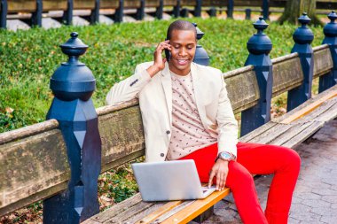 İş adamı dışında. Giyinme krem blazer, yakasız kazak, kırmızı pantolon, kol saati, giyen genç bir adam bankta oturuyor gülümseyen, telefon görüşmesi, dizüstü bilgisayar üzerinde çalışan