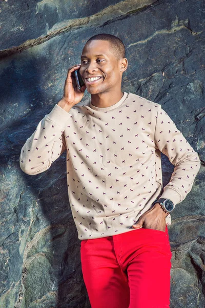 Man Talking on Phone. Dressing in cream, patterned, collarless sweater, red pants, wearing wristwatch, a young black guy is standing by rocks, smiling, making a phone call on his mobile phone