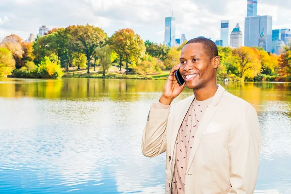 Evet iş adamı. Krem blazer, yakasız kazak, giyinme genç bir siyah adam büyük bir şehirde bir gölün yanında duruyor gülümseyerek, cep telefonundan bir telefon görüşmesi.