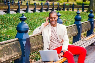 İş adamı dışında. Giyinme krem blazer, yakasız kazak, kırmızı pantolon, kol saati, giyen genç bir adam bankta oturuyor gülümseyen, telefon görüşmesi, dizüstü bilgisayar üzerinde çalışan