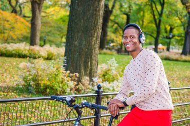 Adam müzik dinleme. Kırmızı pantolon, desenli yakasız süveterin içinde giyinme giyen kulaklık, zenci bir bisiklet Park, devirme bir genç gülümseyen, mutlu bir şekilde bakıyorsun. Sonbahar gezisi.