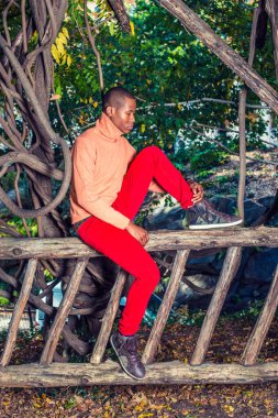 Dışında düşünme. Yüksek yaka, kırmızı pantolon ile ışık turuncu kazaklı giyinme, desenli çizme ayakkabı, siyah adam çit, oturan genç bir bacak bükme, aşağı bakıyor, düşünme, dalıp gitmişim.