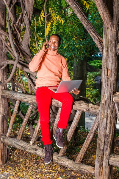 Man Working Outside. Dressing in light orange sweater with high collar, red pants, patterned boot shoes, a young black guy is sitting on fence, smiling, making phone call, working on laptop computer.