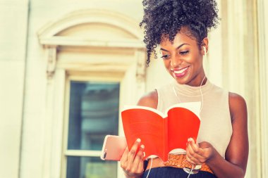 Kulaklık ve cep telefonu, müzik dinleme kırmızı kitap, okuma mutlu Afrikalı-Amerikalı genç bayan üniversite öğrencisi tarafından vintage tarzı ofis binası New York'ta oturan afro saç modeli ile gülümseyen