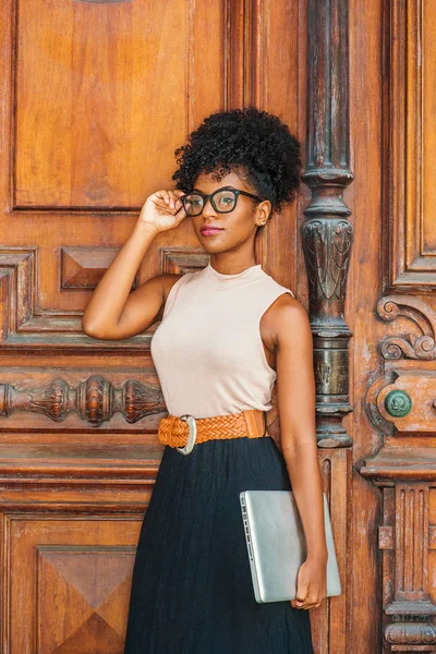 Young African American College Student with afro hairstyle, eye glasses, wearing sleeveless light color top, black skirt, belt, holding laptop computer, standing by vintage office door in New York