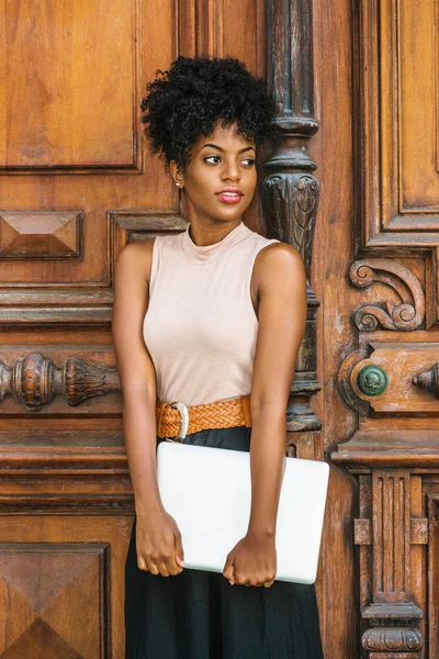 Young African American College Student with afro hairstyle, wearing sleeveless light color top, black skirt, belt, hands holding laptop computer in front, standing by vintage office door in New York