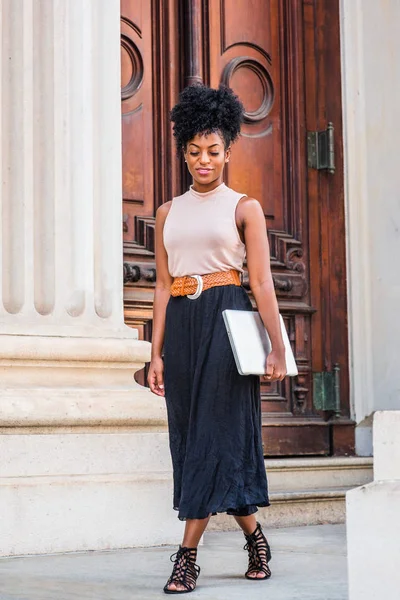 Young black woman with afro hairstyle, wearing sleeveless light color top, black skirt, belt, strappy sandals, carrying laptop computer, walking by office doorway in New York, looking down, thinking