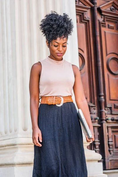 Young black woman with afro hairstyle, wearing sleeveless light color top, black skirt, dark orange belt, holding laptop computer, standing by office doorway in New York, looking down, thinking
