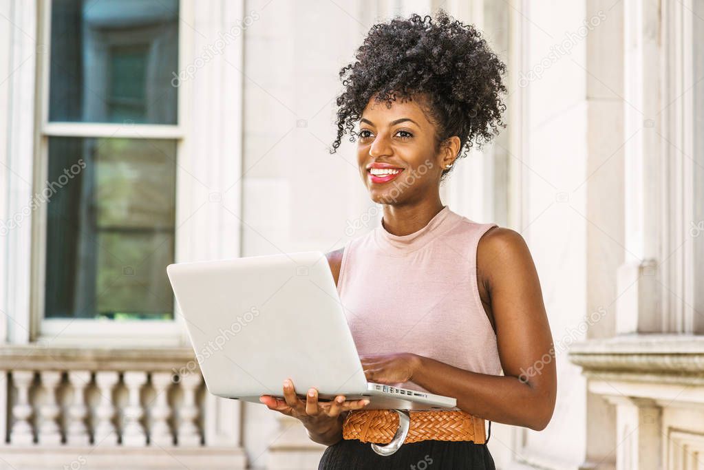 Young African American woman with afro hairstyle wearing sleeveless light color top, belt, standing in vintage office building in New York, working on laptop computer, looking, thinking, smiling