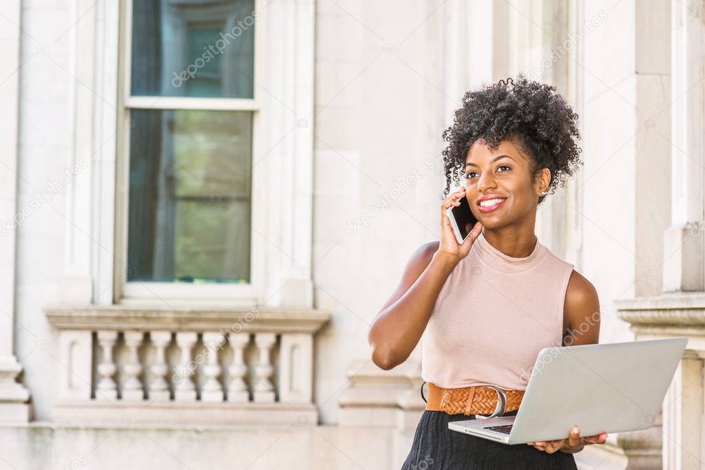 African American Businesswoman working in New York. Young black college student with afro hairstyle sitting on railing in vintage style office building, working on laptop computer, making phone call.