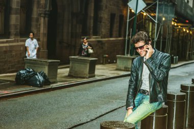 Siyah deri ceket, kot, kahverengi çizme ayakkabı, güneş gözlüğü, New York, sokakta oturan sakallı genç bir adam takıyor gülümseyen, telefonda konuşurken, rahatlatıcı.