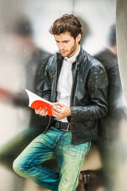 Okuma gücünü. Siyah deri ceket, beyaz gömlek, gümüş metal duvara yaslanmış giyen genç adam sakallı, eller kırmızı kitap, aşağı bakıyor, ara çalışma sırasında okuma.